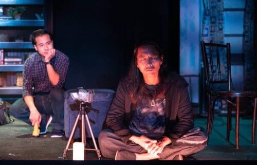 Two people sitting on stage in a dimly lit setting, one speaking into a microphone.