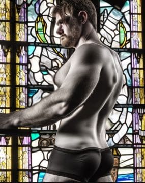 Muscular shirtless man posing in front of a stained glass window.