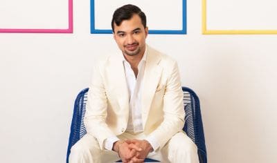 Man in white suit sitting on a blue chair smiling.