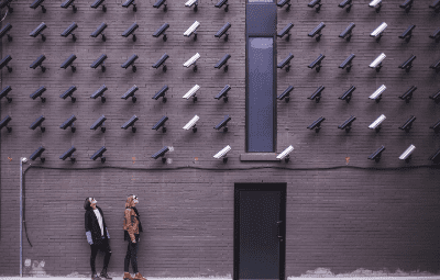 Two people walk past a wall covered with numerous security cameras.