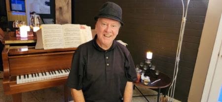 Smiling elderly man in black outfit and cap near piano.