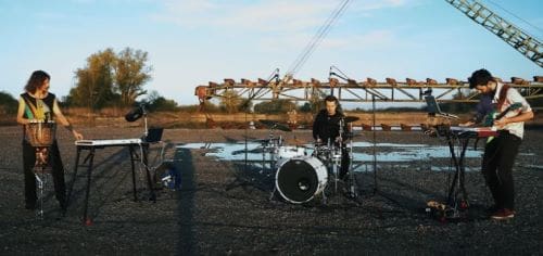 Musicians playing drums outdoors near water at sunset.