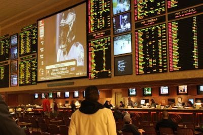Person watching live betting odds and sports on large screens inside a sportsbook.