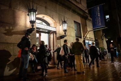People queue outside a warmly lit building at night.