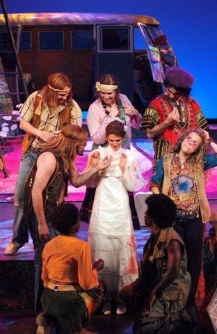 Actors in tribal costumes perform on stage with a central figure in white.