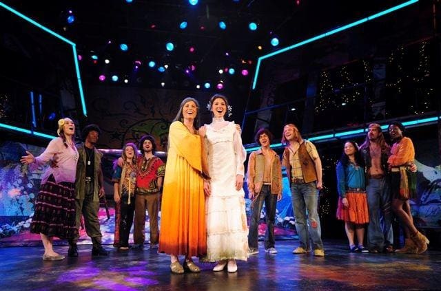 Two women stand center stage in elegant dresses amid a colorful theatrical cast.