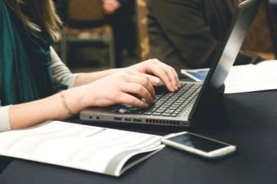 A person typing on a laptop with an open book and smartphone nearby.