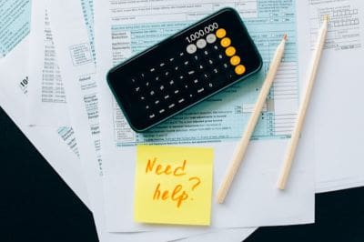 Calculator, pens, and a sticky note saying 'Need help?' on a newspaper.