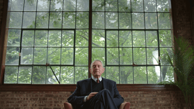 An elderly man in a suit sits thoughtfully by a large window with greenery outside.