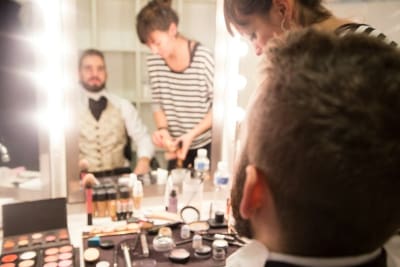 A man getting his makeup done in a busy dressing room.