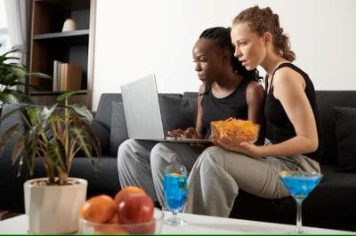 Two friends enjoying snacks while watching something on a laptop.