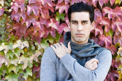Man in gray sweater and scarf stands before red and green ivy-covered wall.