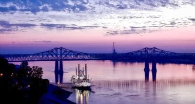 A steamboat sails under a bridge at sunset with a colorful sky.