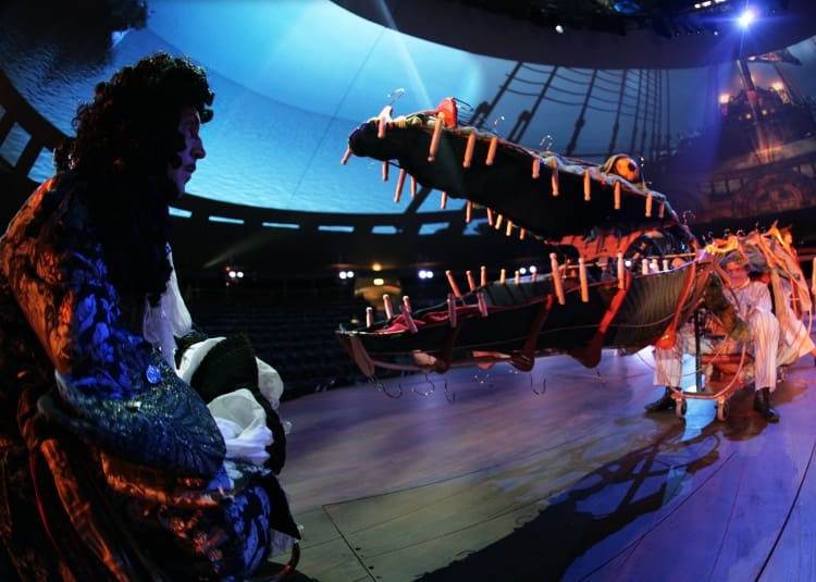A person faces a large crocodile with candles on its teeth in a dramatic setting.