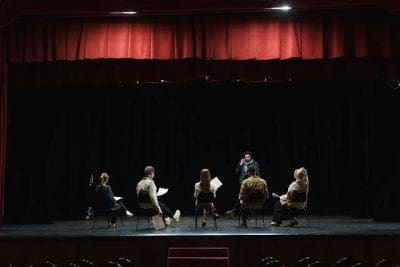 A group of six people seated in a semi-circle on a stage with a red curtain backdrop.