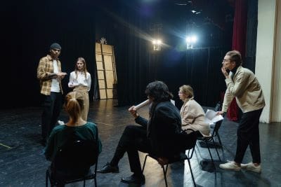 Actors rehearsing on a dimly lit stage with chairs.