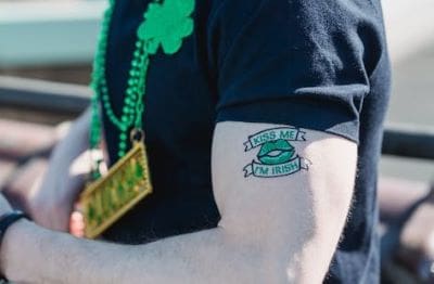 A person wearing a black shirt with a frog tattoo on their arm and a green shamrock necklace.