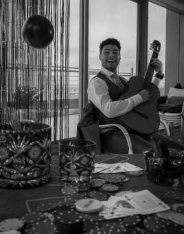 A man playing guitar, smiling in a cozy room with candles and decorations.