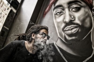 Man exhales smoke near a mural of Tupac Shakur.