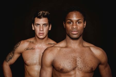 Two shirtless men standing closely together against a dark background.