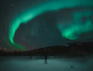 Person standing under vibrant green northern lights in a snowy landscape.