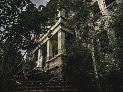 An old, abandoned building surrounded by overgrown vegetation and shadowy trees.