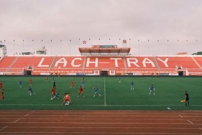 Football match in progress at Lach Tray stadium with players on the field and empty stands.