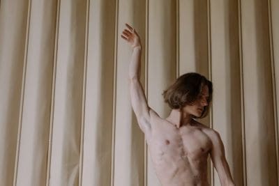 Shirtless person with raised arm against a beige curtain background.