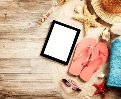 A tablet, pink flip-flops, sunglasses, and a sunhat on wooden surface.