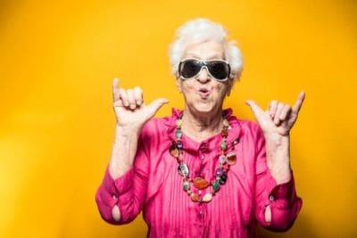 Stylish elderly woman in sunglasses and pink outfit posing against a yellow background.