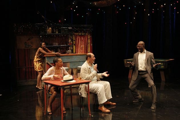 Actors performing a dramatic scene on stage with a rustic backdrop.