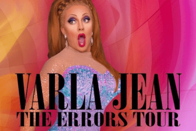 Drag queen in vibrant makeup and costume on a pink stage backdrop.