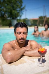 Young man relaxing by the pool with a drink, others socializing in the background.
