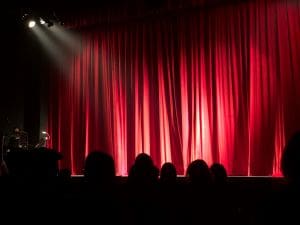 A red theater curtain illuminated by stage lights with a dark audience in front.