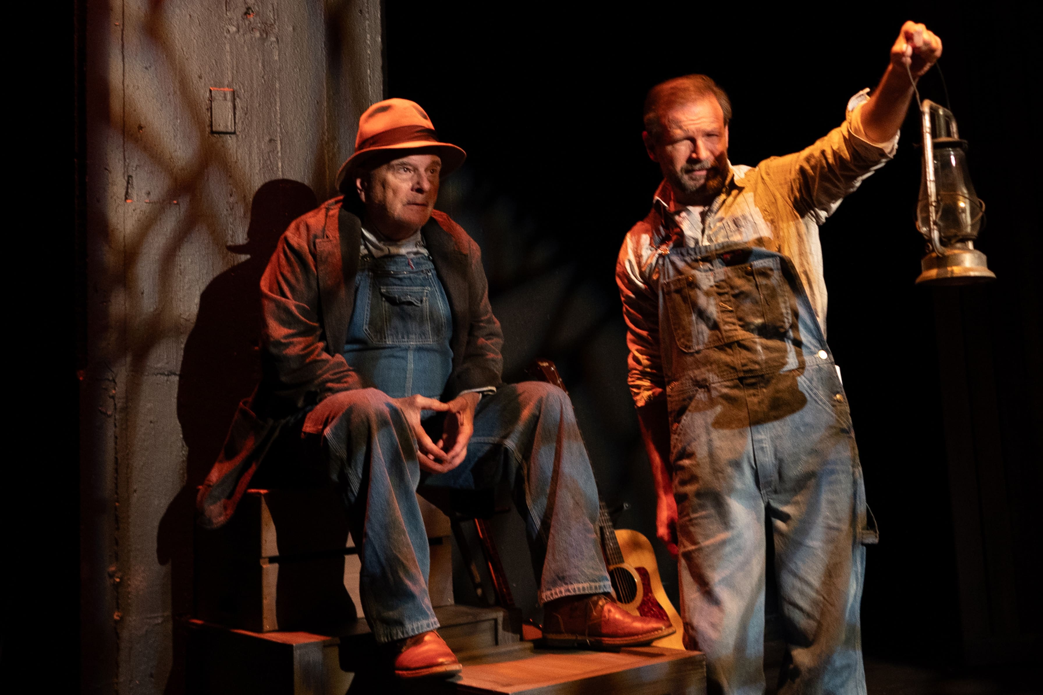 Two actors in work clothes performing on stage with dramatic lighting.