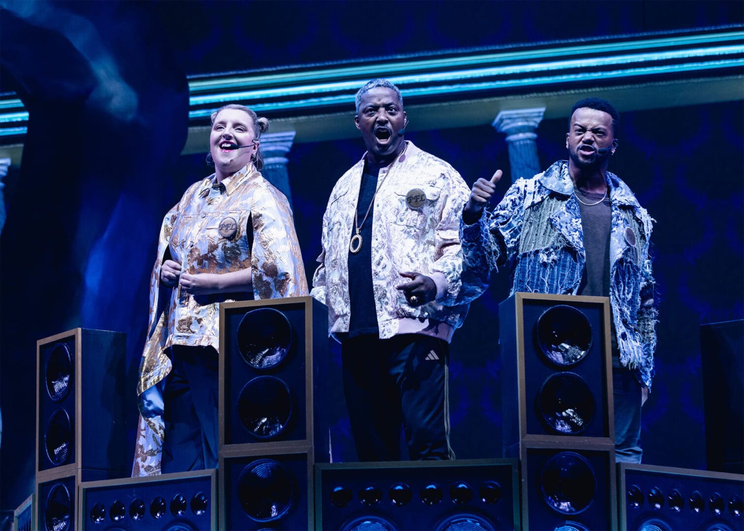 Three performers on stage with bright costumes and speakers.