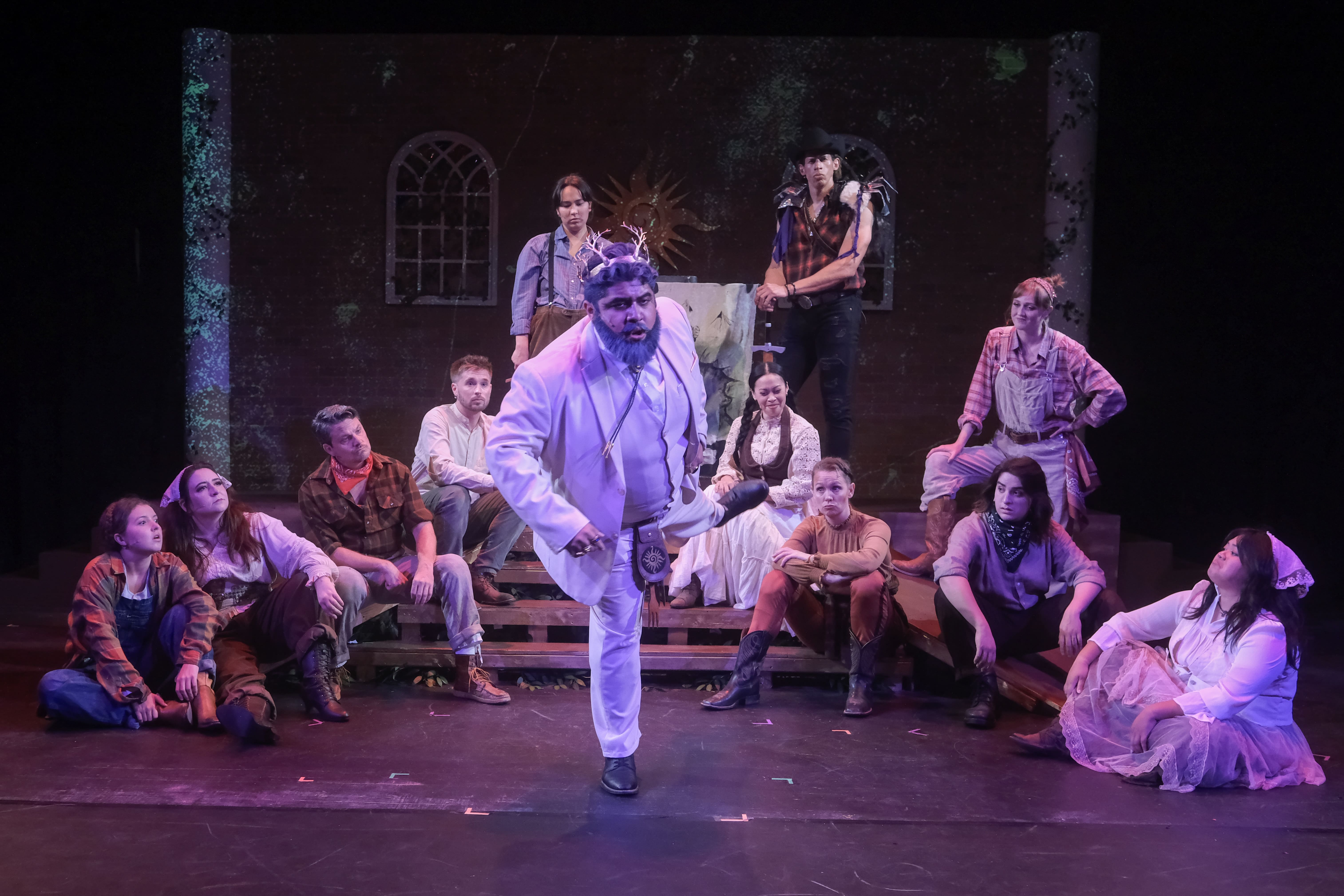 A man in white dances energetically on stage with a lively cast behind him.