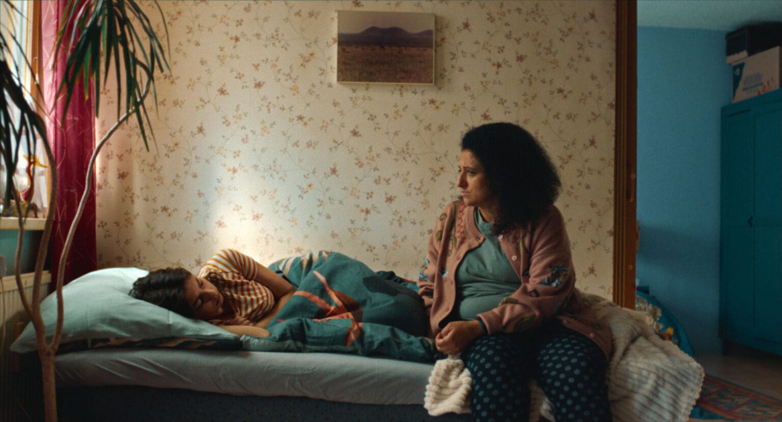 A woman sits pensively beside a sleeping child in a dimly lit room.