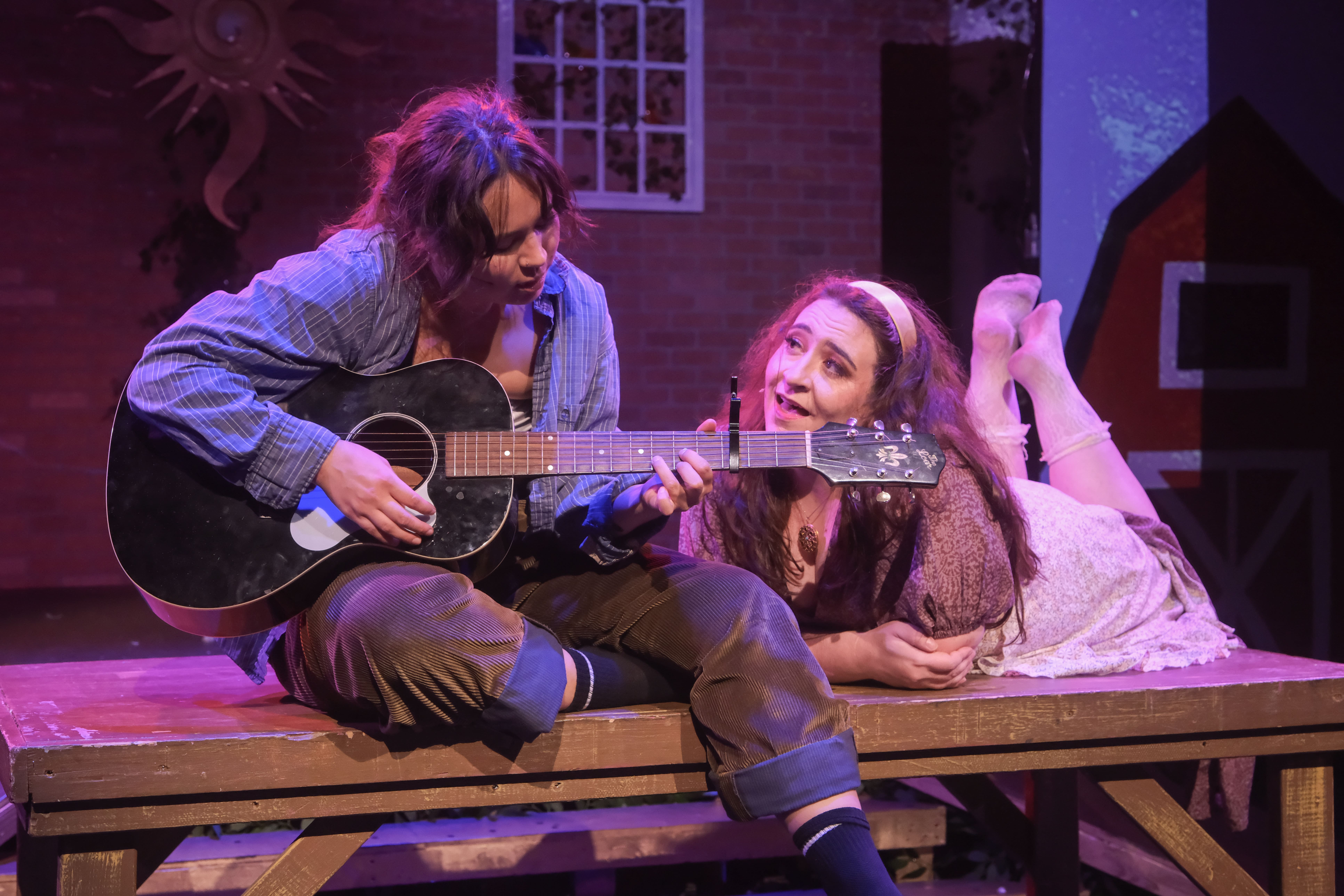 Man playing guitar to a woman lying down, both on a wooden stage.