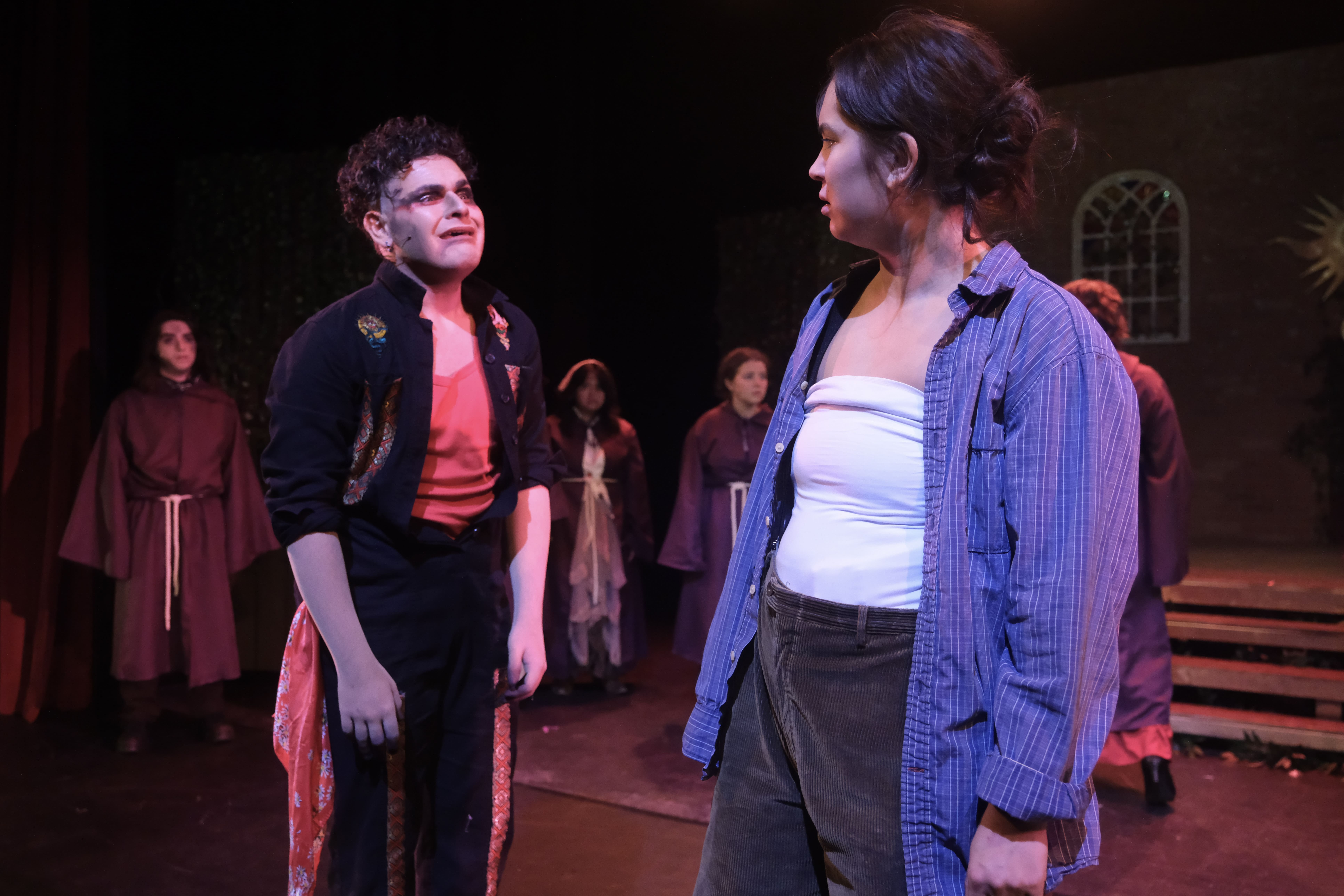 Two young actors engage in a tense scene on stage with others in the background.