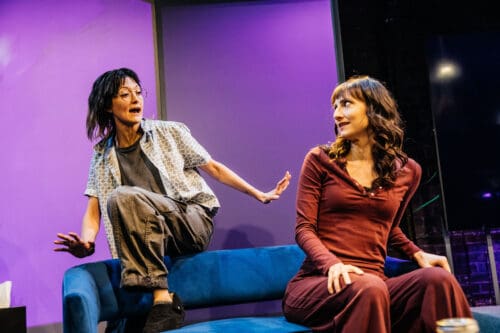 Two women engaged in an intense conversation on stage with dramatic lighting.