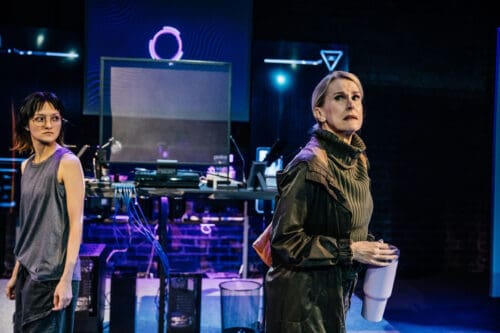 Person in futuristic attire standing near neon-lit equipment at night.
