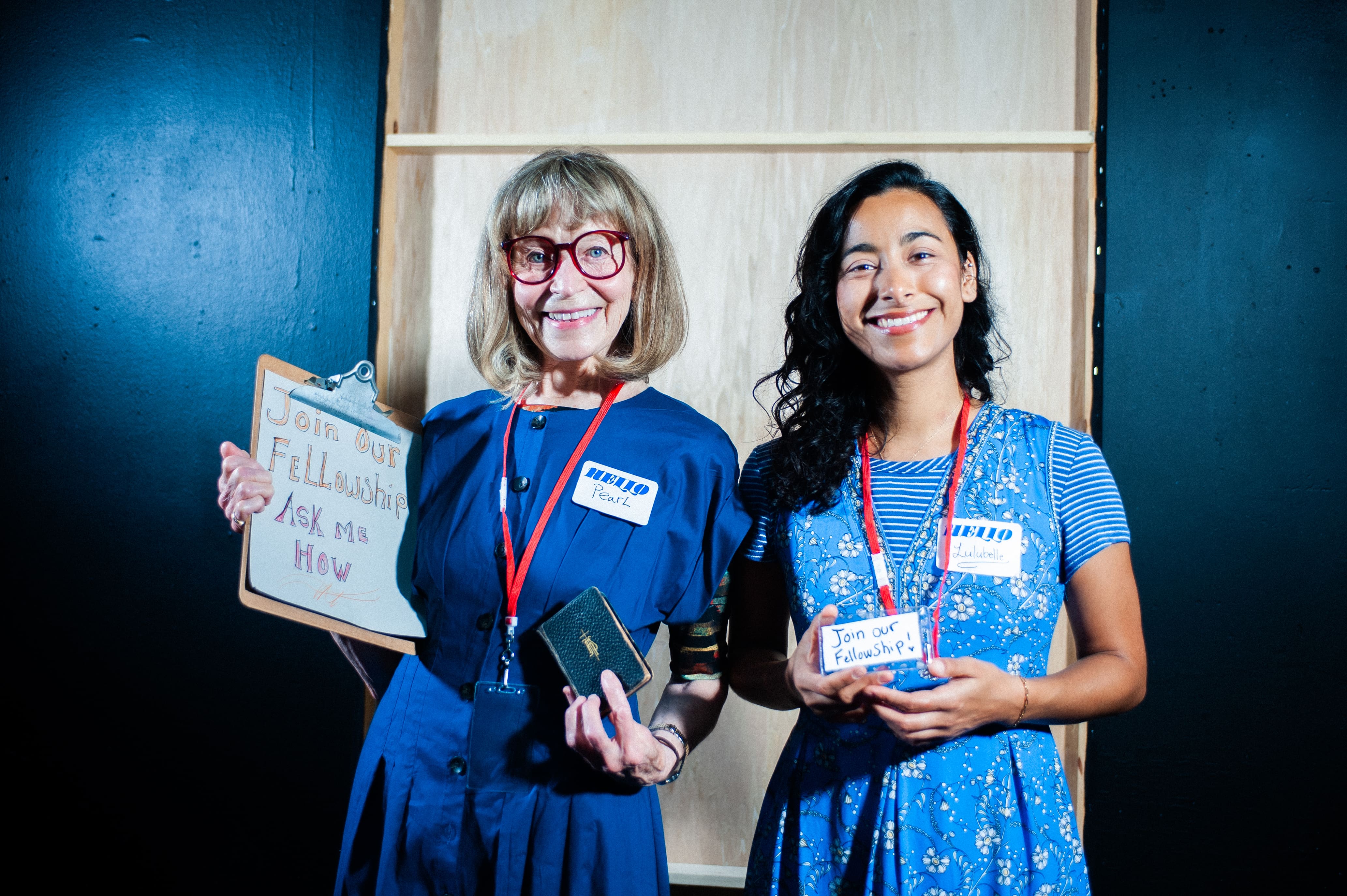 Two women smiling, wearing blue dresses and name tags, holding a phone and a notebook.