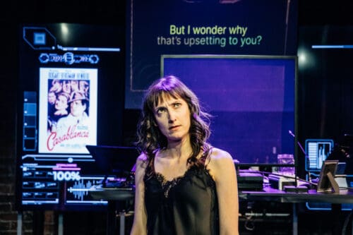 A woman stands under purple lighting with a neon sign behind her.