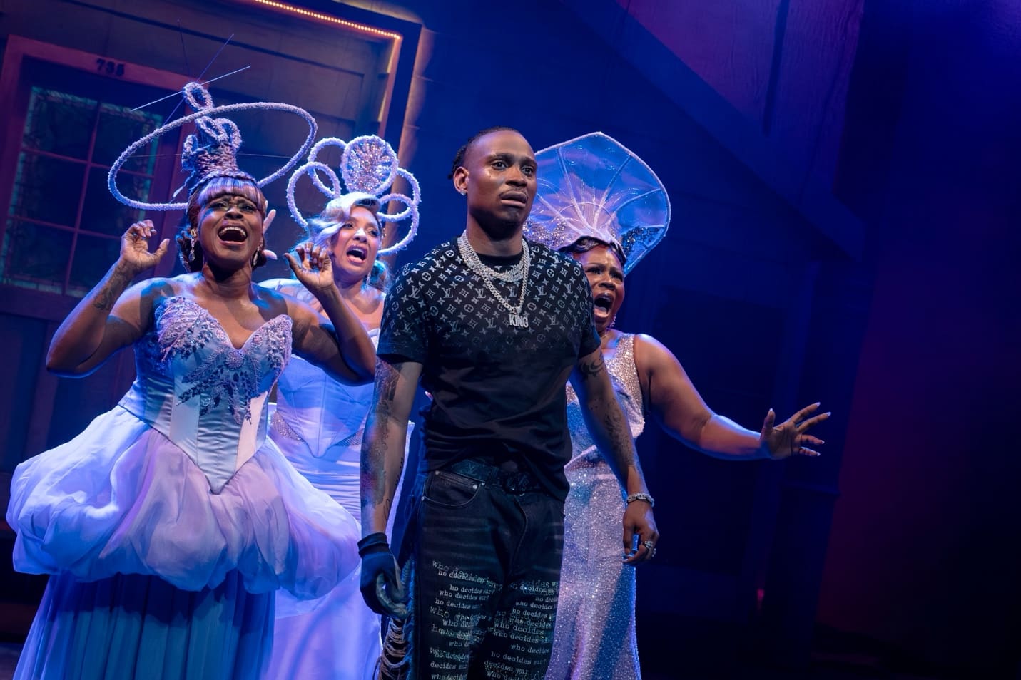 A man stands confidently on stage with performers in whimsical costumes behind him.