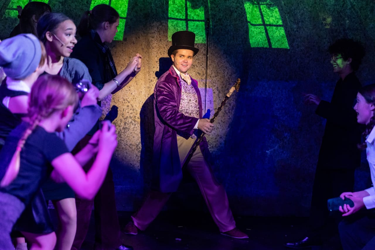 A performer in a purple suit and top hat holding a cane on stage.