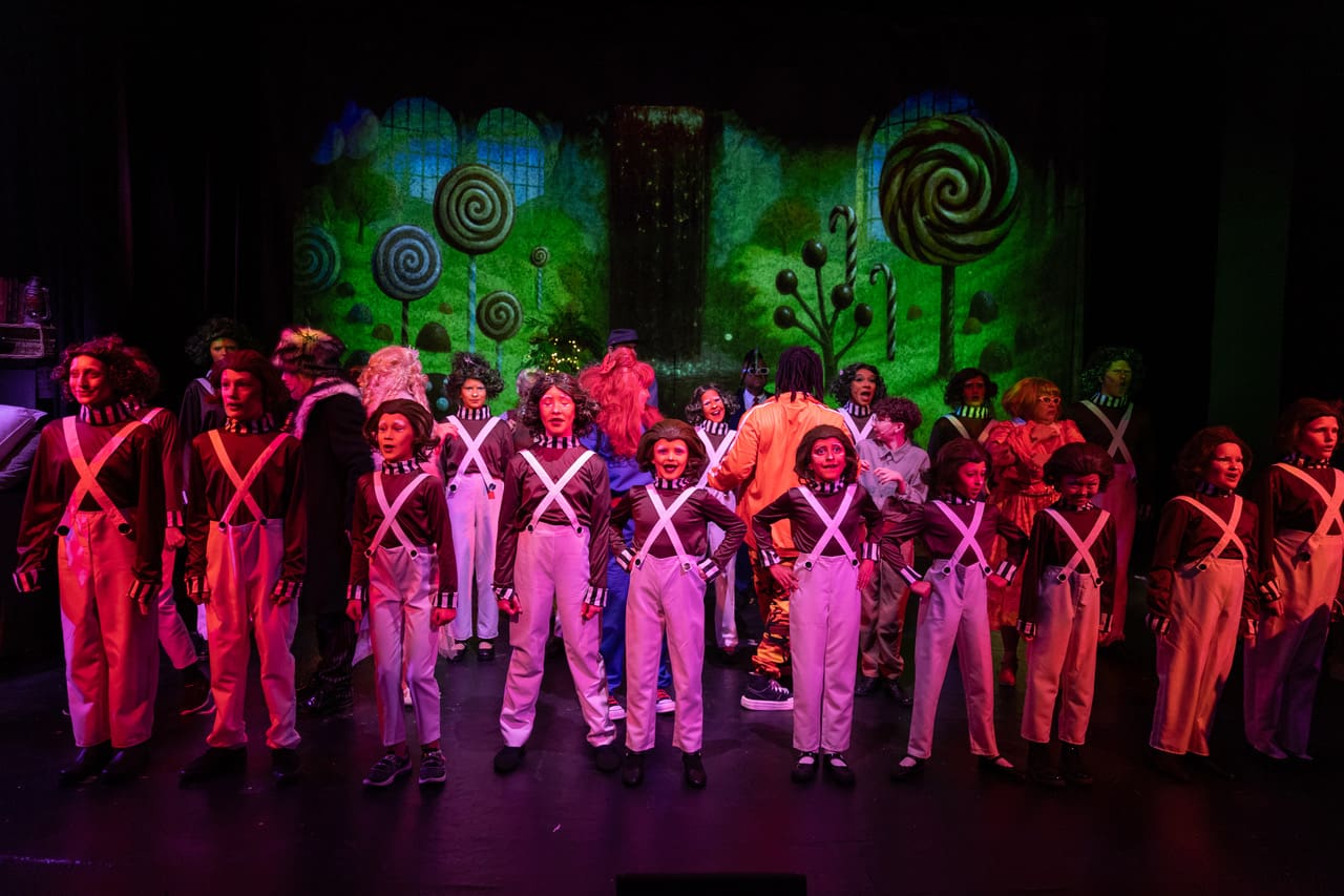 Actors dressed as Oompa Loompas perform on stage in a colorful set.