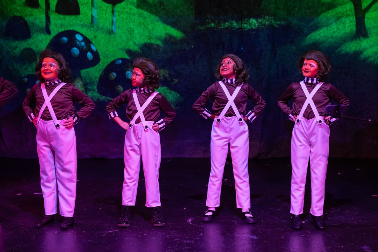 Four performers in white outfits and suspenders stand on stage with colorful backdrop.