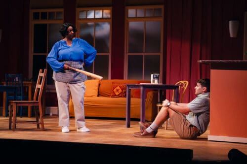 Two actors engage in a dramatic scene on a cozy stage set with a sofa and coffee table.