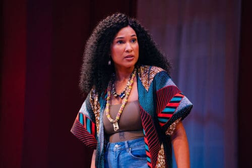 A woman with curly hair wearing a colorful cardigan and layered necklaces.